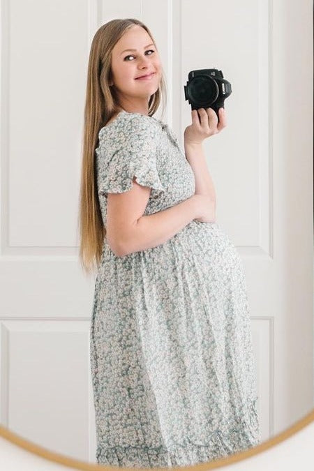 woman wearing a floral maternity dress and holding her baby bump while looking at the camera