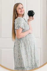 woman wearing a floral maternity dress and holding her baby bump while looking at the camera