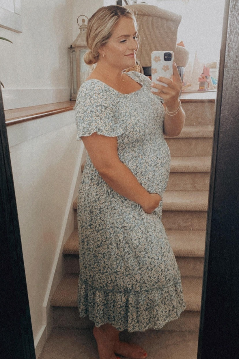 woman wearing a light blue floral maternity dress, gently holding her baby bump while standing by a staircase