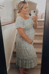 woman wearing a light blue floral maternity dress, gently holding her baby bump while standing by a staircase