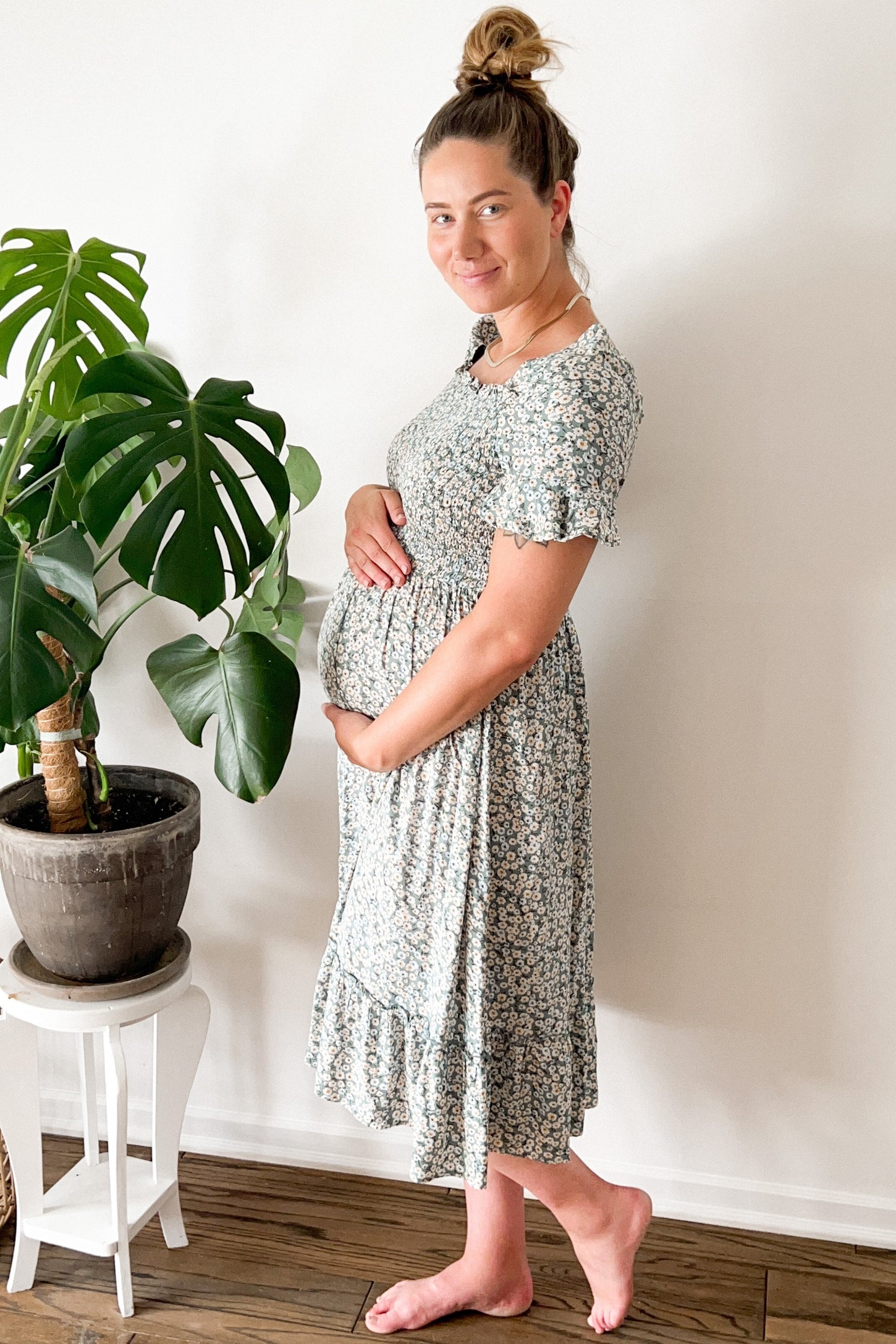 woman wearing a floral maternity dress, cradling her baby bump, standing next to a potted plant