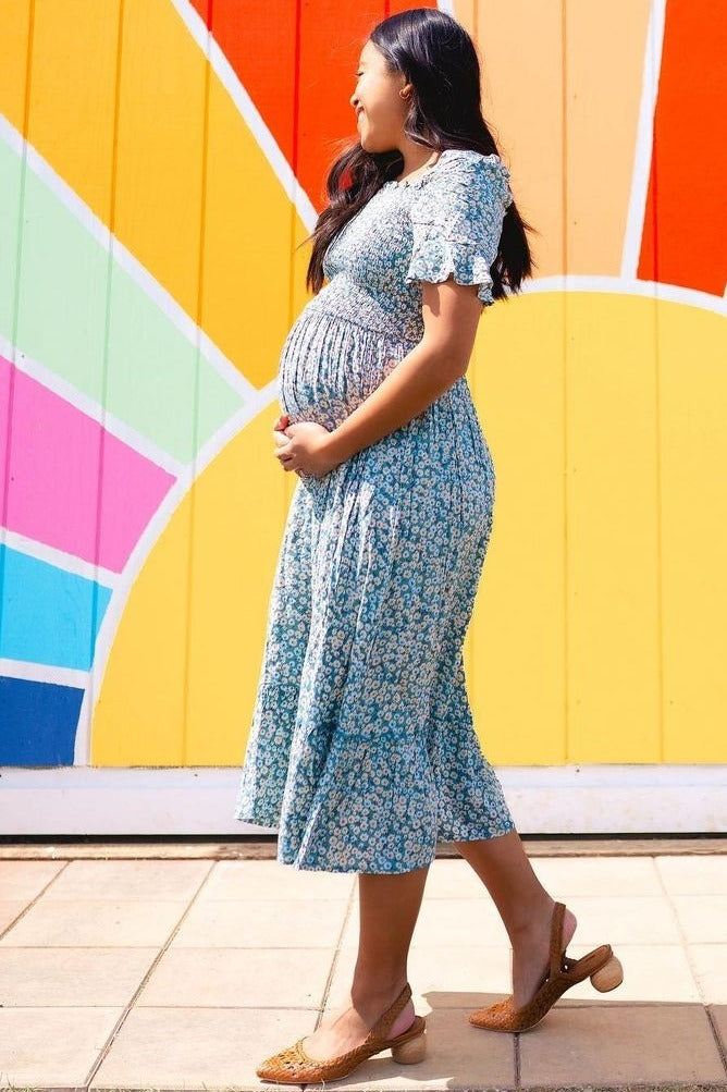 woman wearing a blue floral print maternity dress and holding her baby bump while walking outdoors