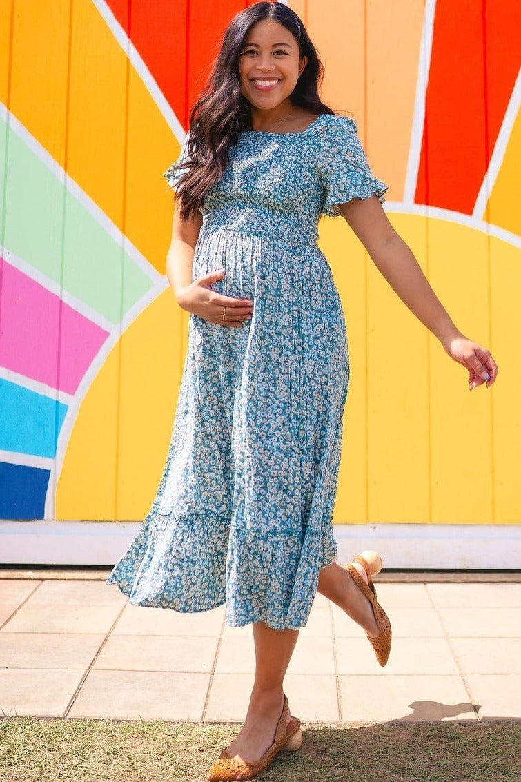 woman wearing a blue floral maternity dress and holding her baby bump, standing in front of a colorful mural