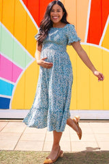 woman wearing a blue floral maternity dress and holding her baby bump, standing in front of a colorful mural