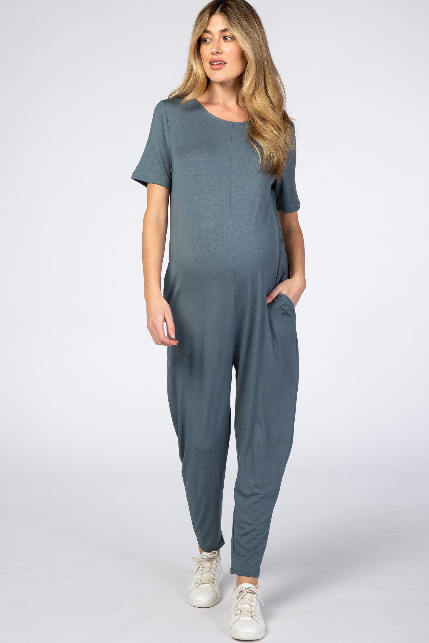 woman wearing a teal short sleeve maternity jumpsuit with side pockets and a visible baby bump