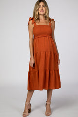 woman wearing an rust orange maternity dress with smocked bodice and tiered skirt, styled with bow straps and a visible baby bump
