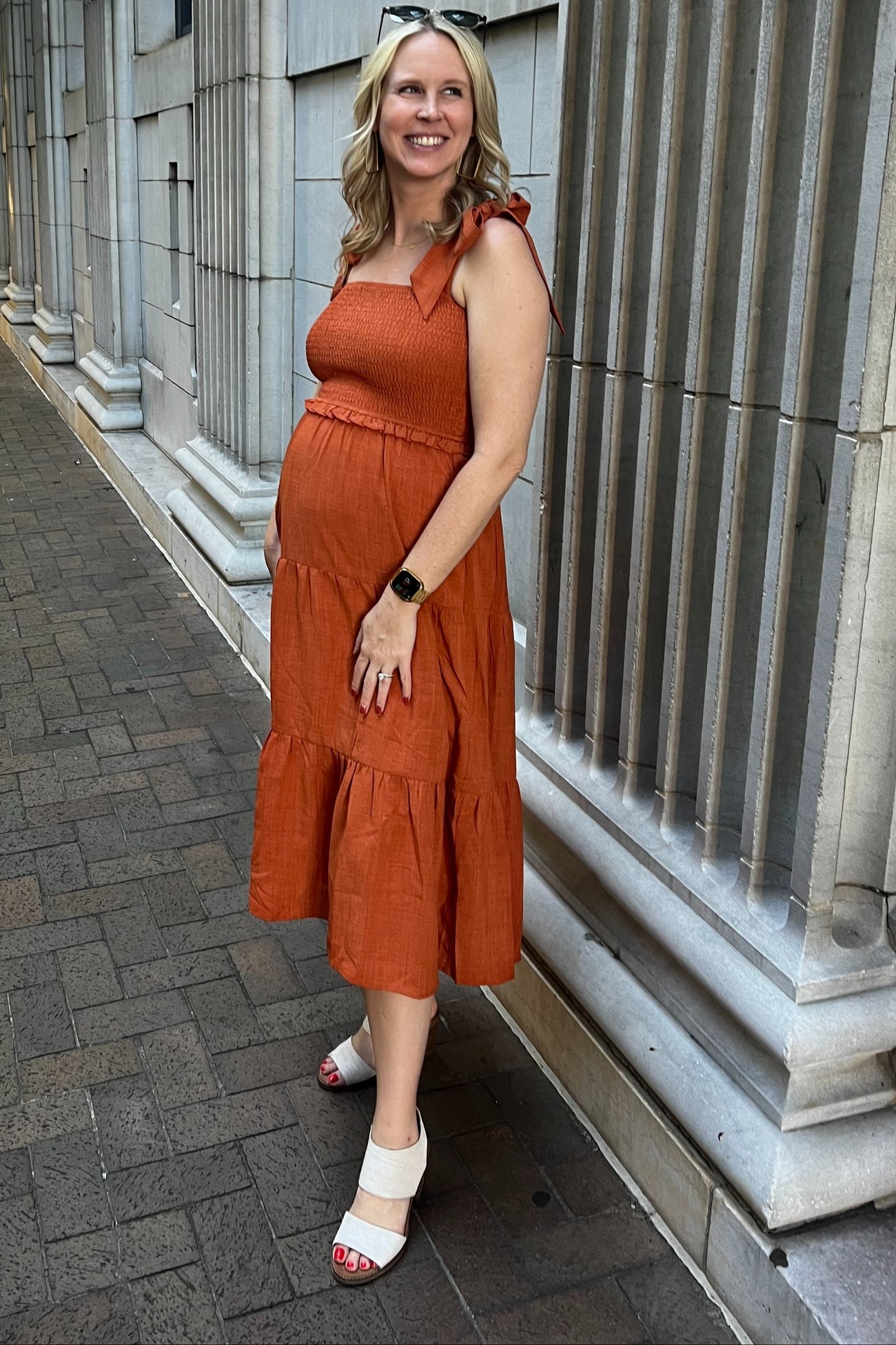 woman wearing an orange maternity sundress with ruffled tiers and a baby bump, standing against a stone wall