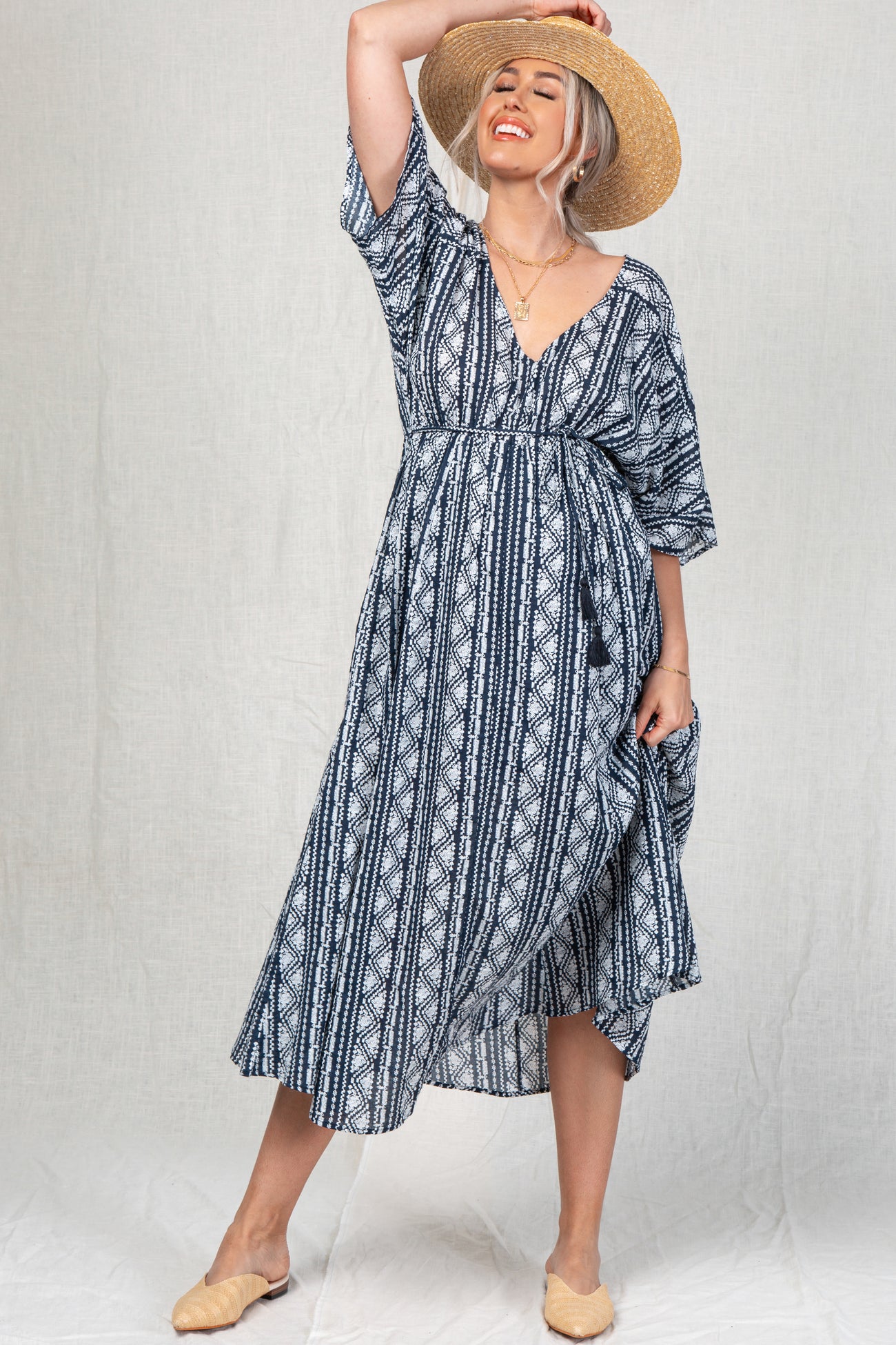 woman wearing a navy and white patterned maternity dress with a v-neck and short sleeves, smiling and holding a straw hat