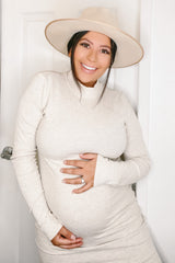smiling pregnant woman in a cream turtleneck dress and wide-brimmed hat holding her baby bump