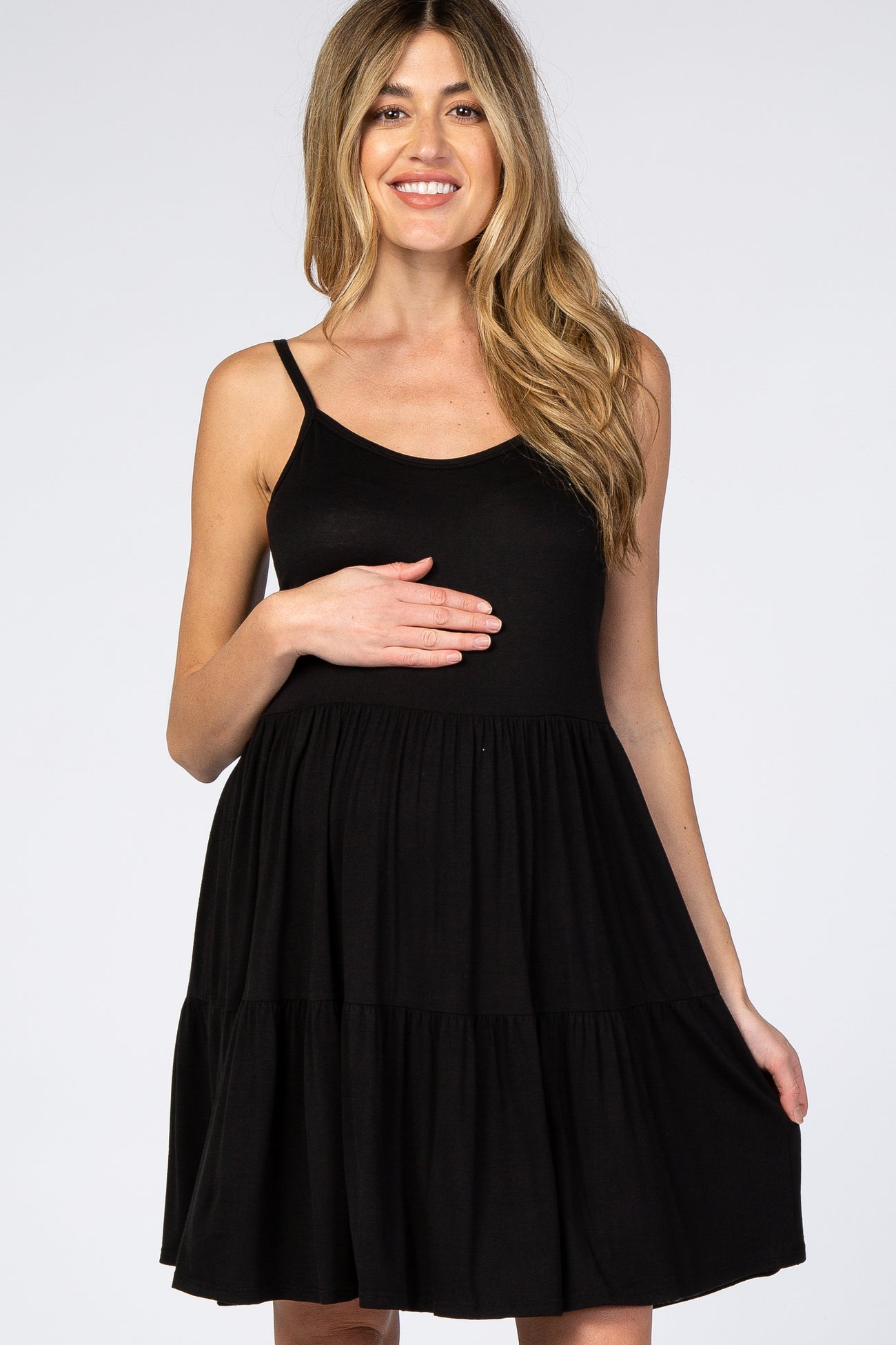 Smiling pregnant woman wearing a black sleeveless tiered maternity dress against a white background