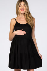 Smiling pregnant woman wearing a black sleeveless tiered maternity dress against a white background