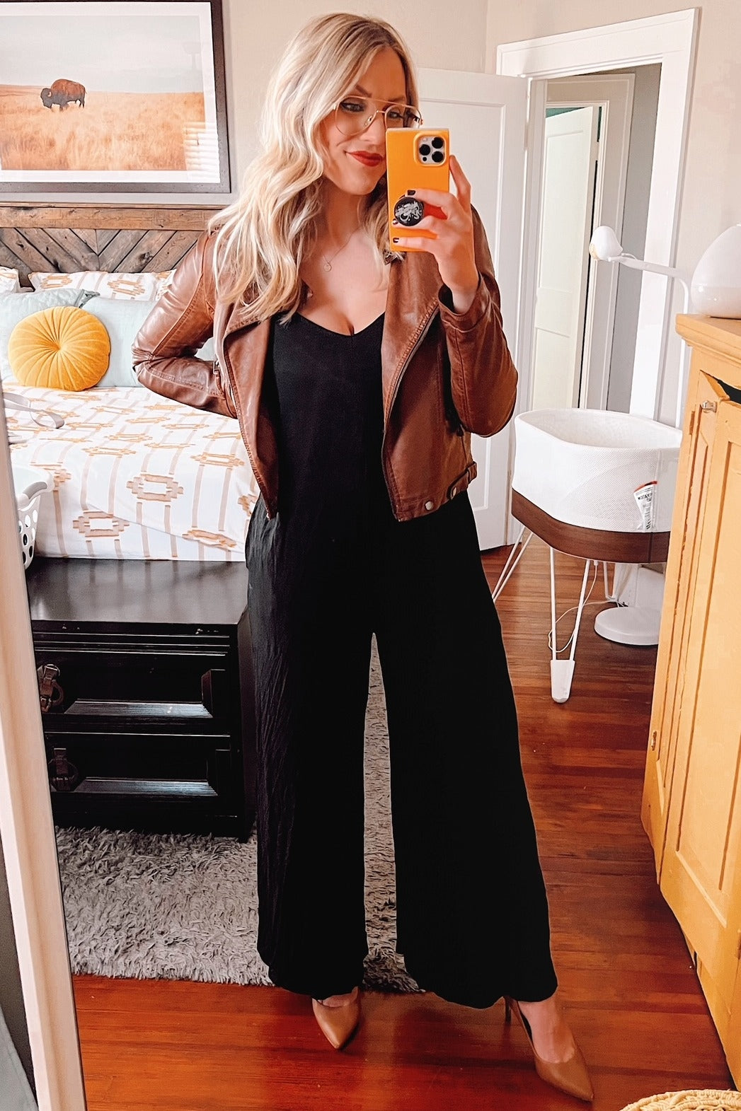 woman wearing a black jumpsuit with a brown leather jacket and tan heels, posing in front of a mirror