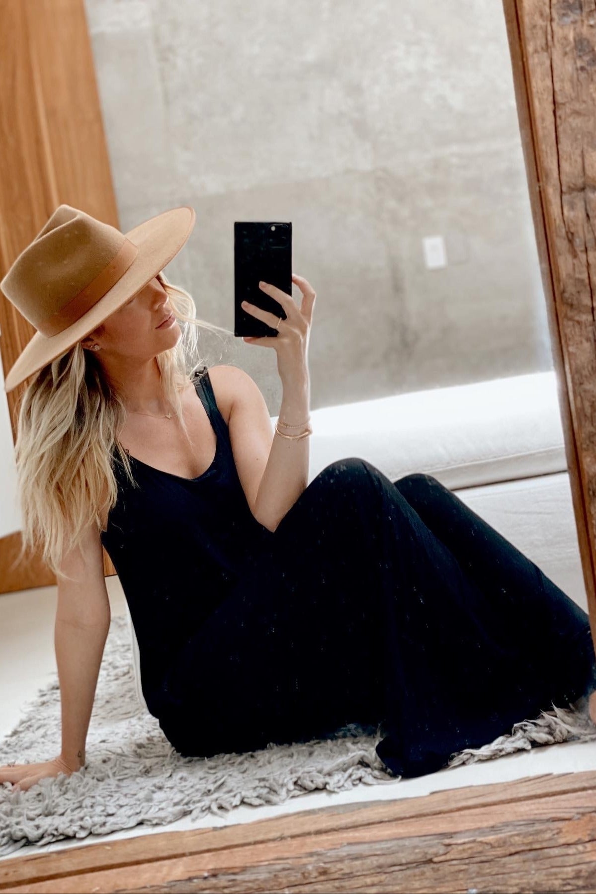 woman wearing a black sleeveless maxi dress and a wide-brimmed tan hat, sitting on the floor in front of a mirror