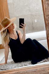 woman wearing a black sleeveless maxi dress and a wide-brimmed tan hat, sitting on the floor in front of a mirror