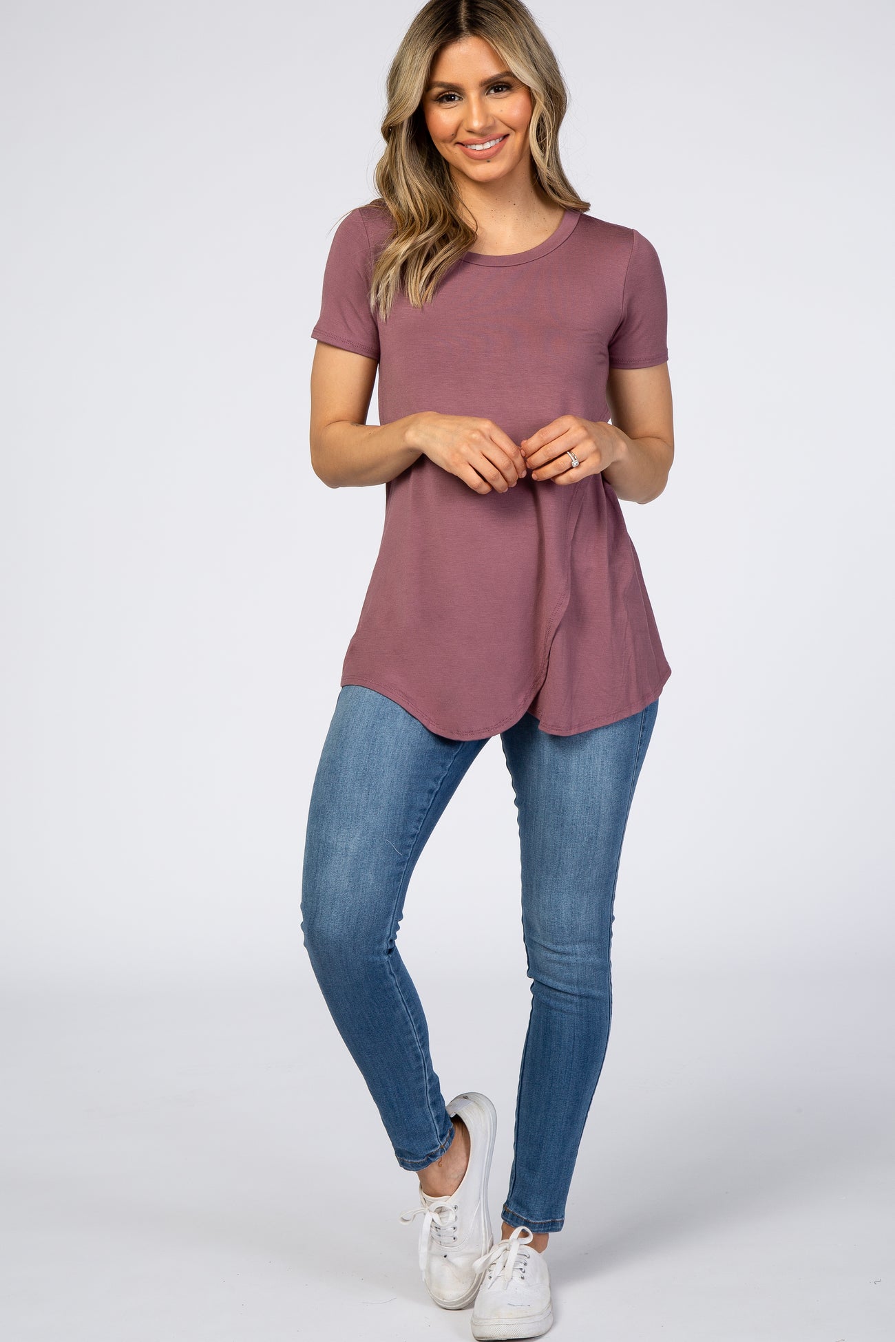 woman wearing a mauve short sleeve top with a curved hem and blue skinny jeans, standing against a plain background
