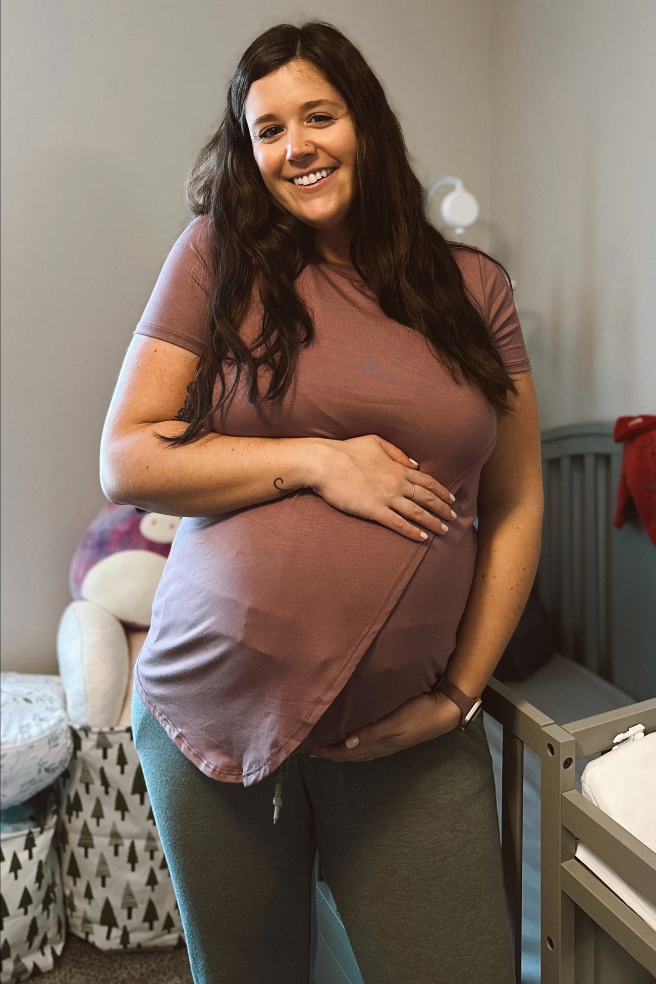 woman wearing a dusty rose maternity t-shirt and gray maternity pants, holding her baby bump in a nursery setting
