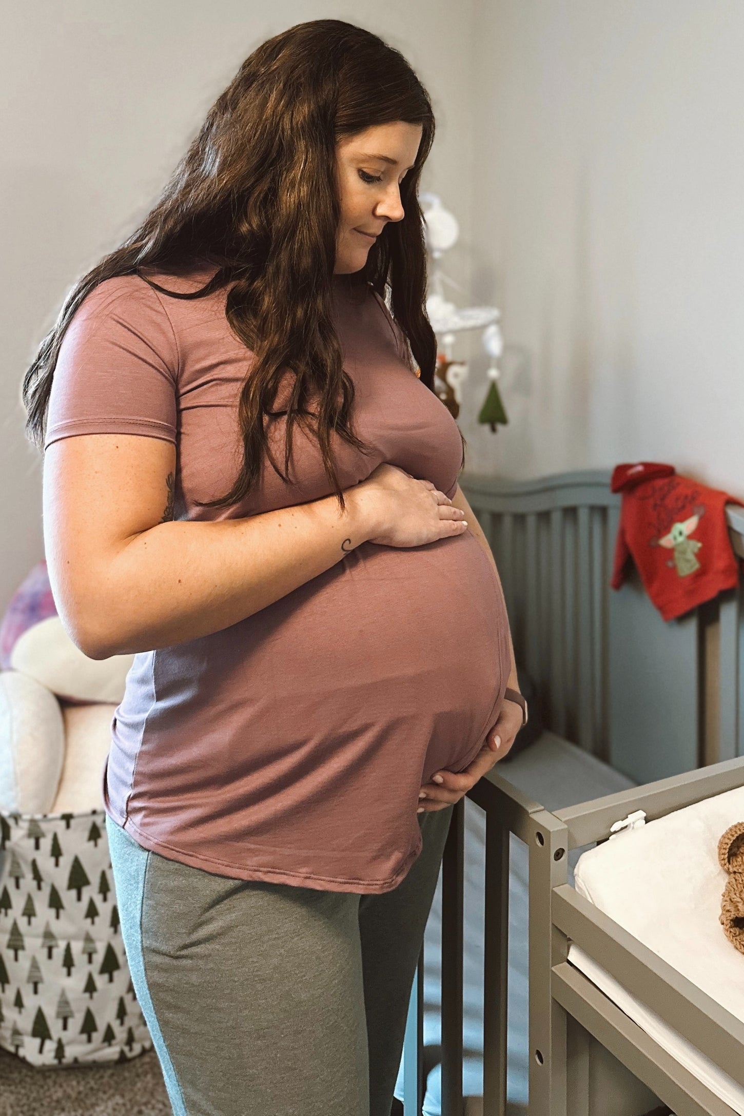 woman wearing a mauve short sleeve maternity top and gray leggings, holding her baby bump in a nursery setting