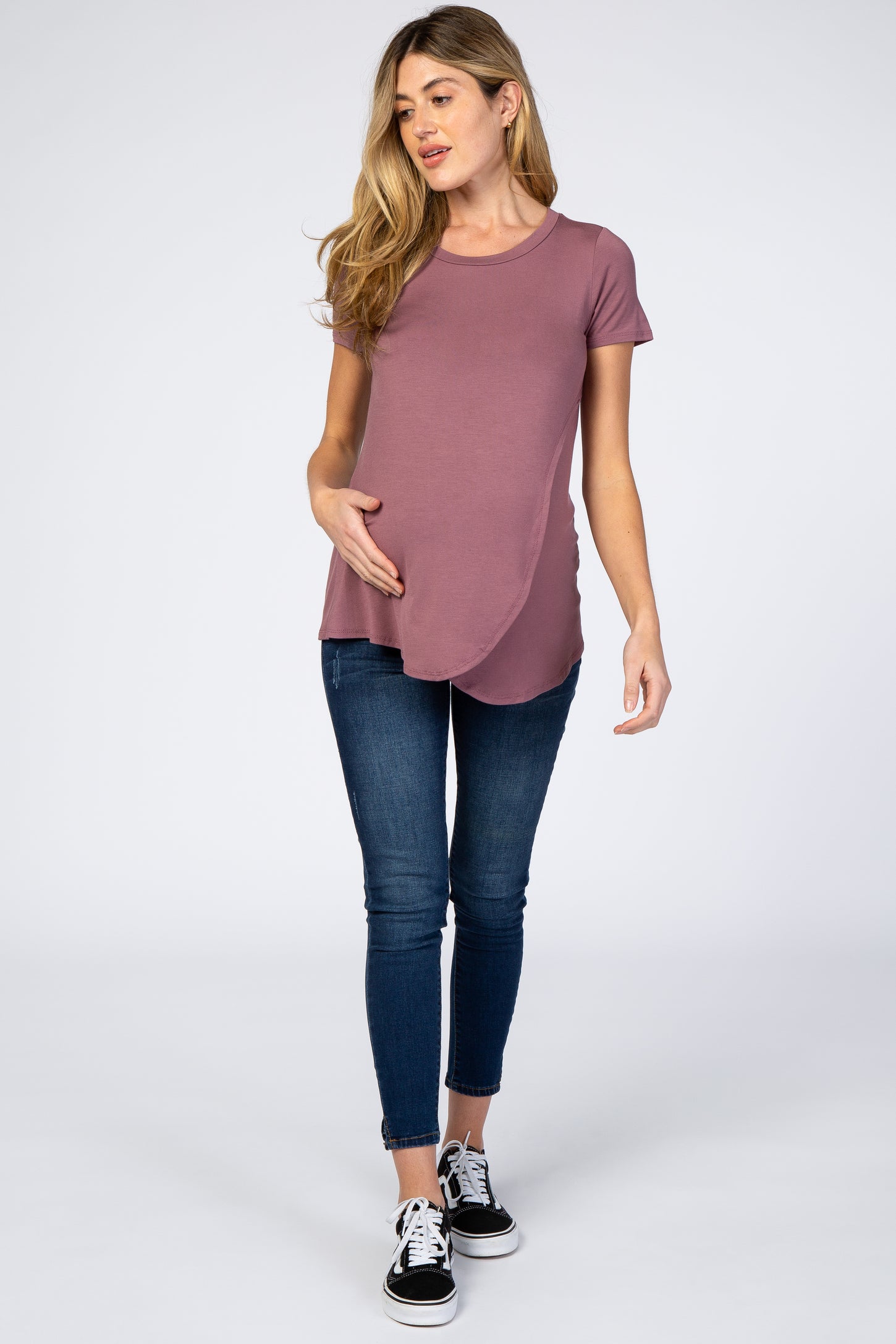 woman wearing a mauve short sleeve maternity top with a curved hem and holding her baby bump, paired with skinny jeans