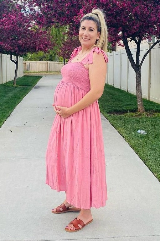 woman wearing a pink maternity dress with tie straps and holding her baby bump in a park setting