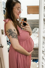 woman wearing a pink maternity dress with shoulder ties and holding her baby bump in front of a mirror