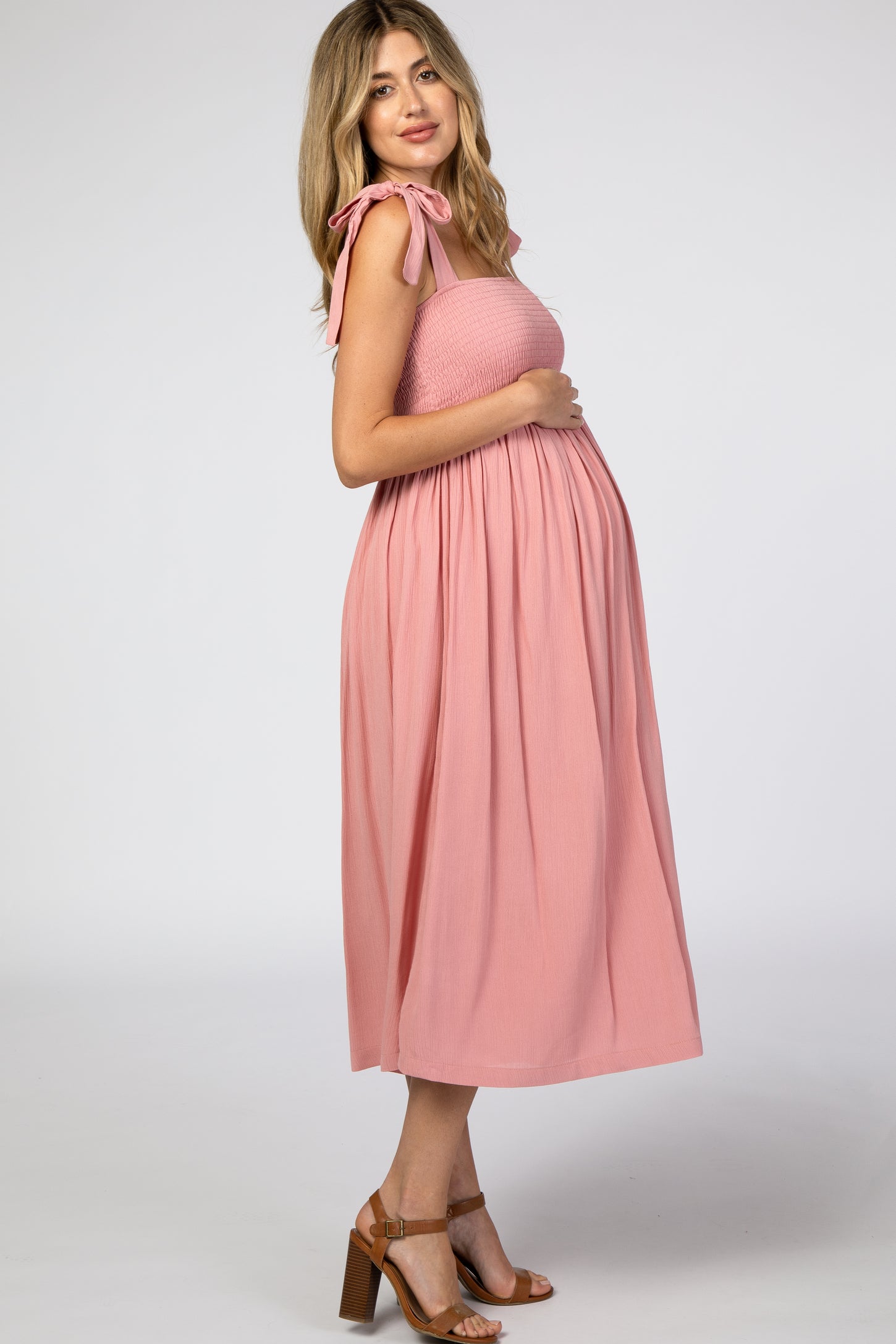 woman wearing a pink smocked maternity dress with tie straps and holding her baby bump
