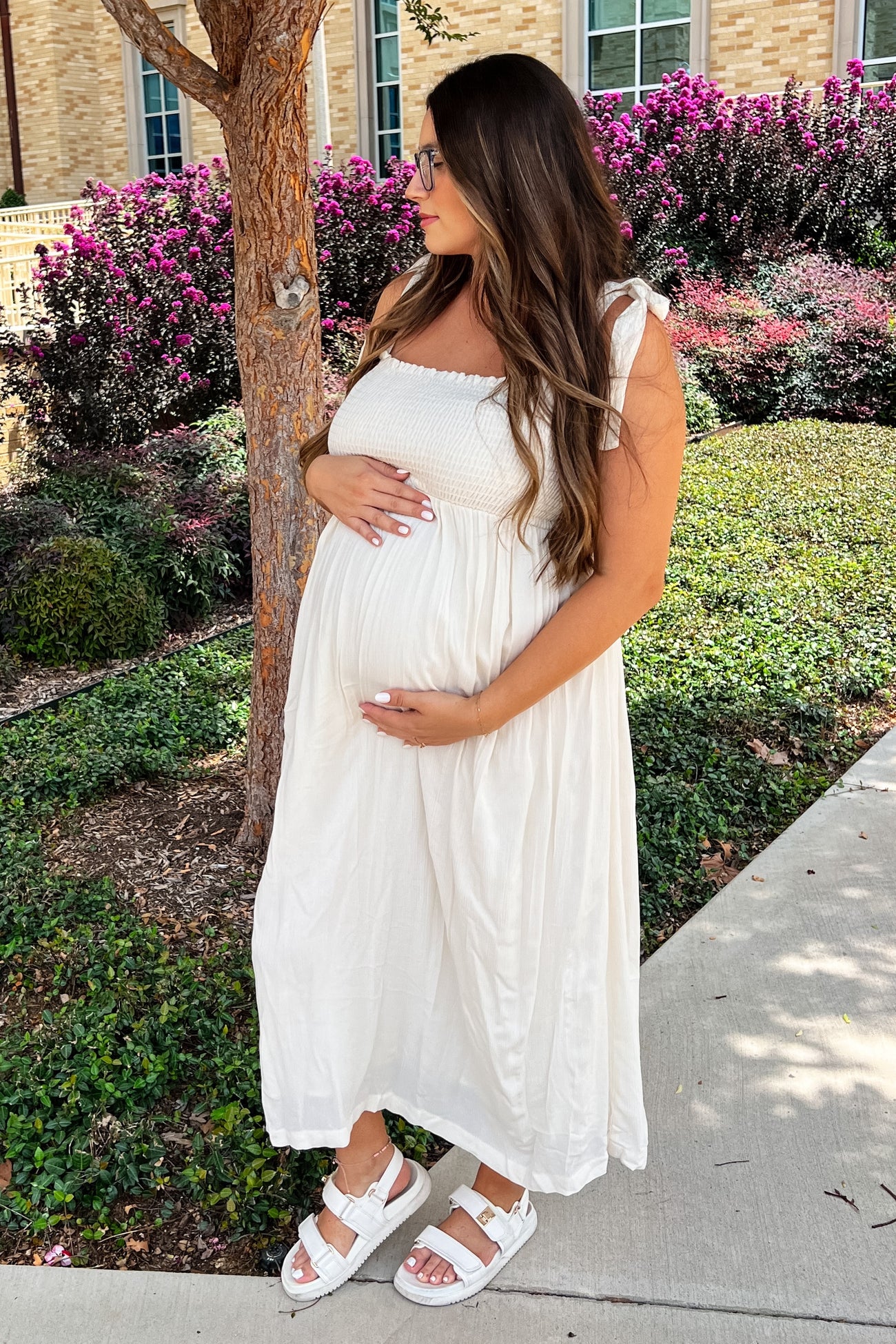 woman wearing a cream maternity dress with smocking detail and adjustable straps, holding her baby bump.