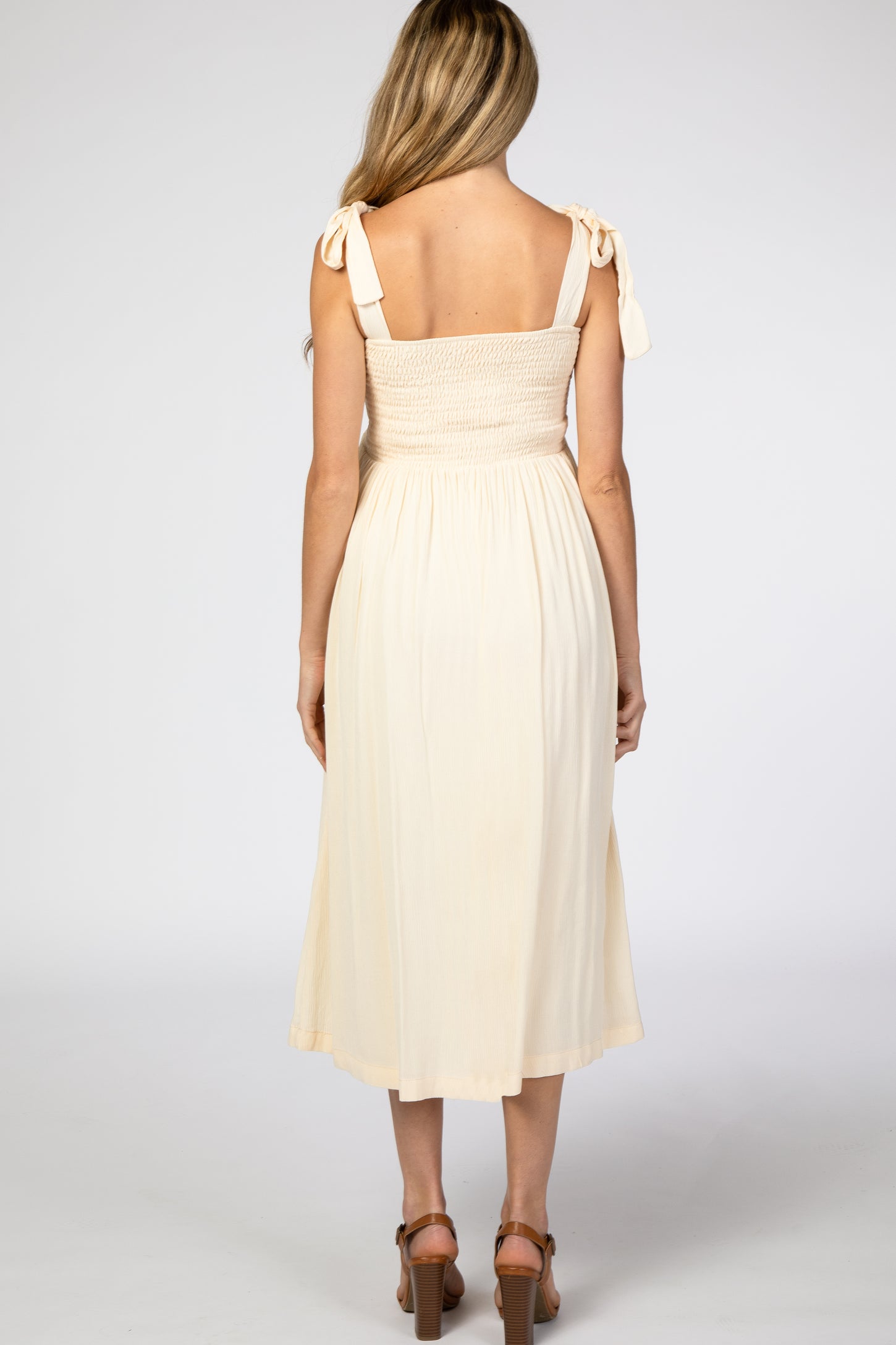 woman wearing a light beige maternity dress with shoulder ties and smocked bodice, showing the back view