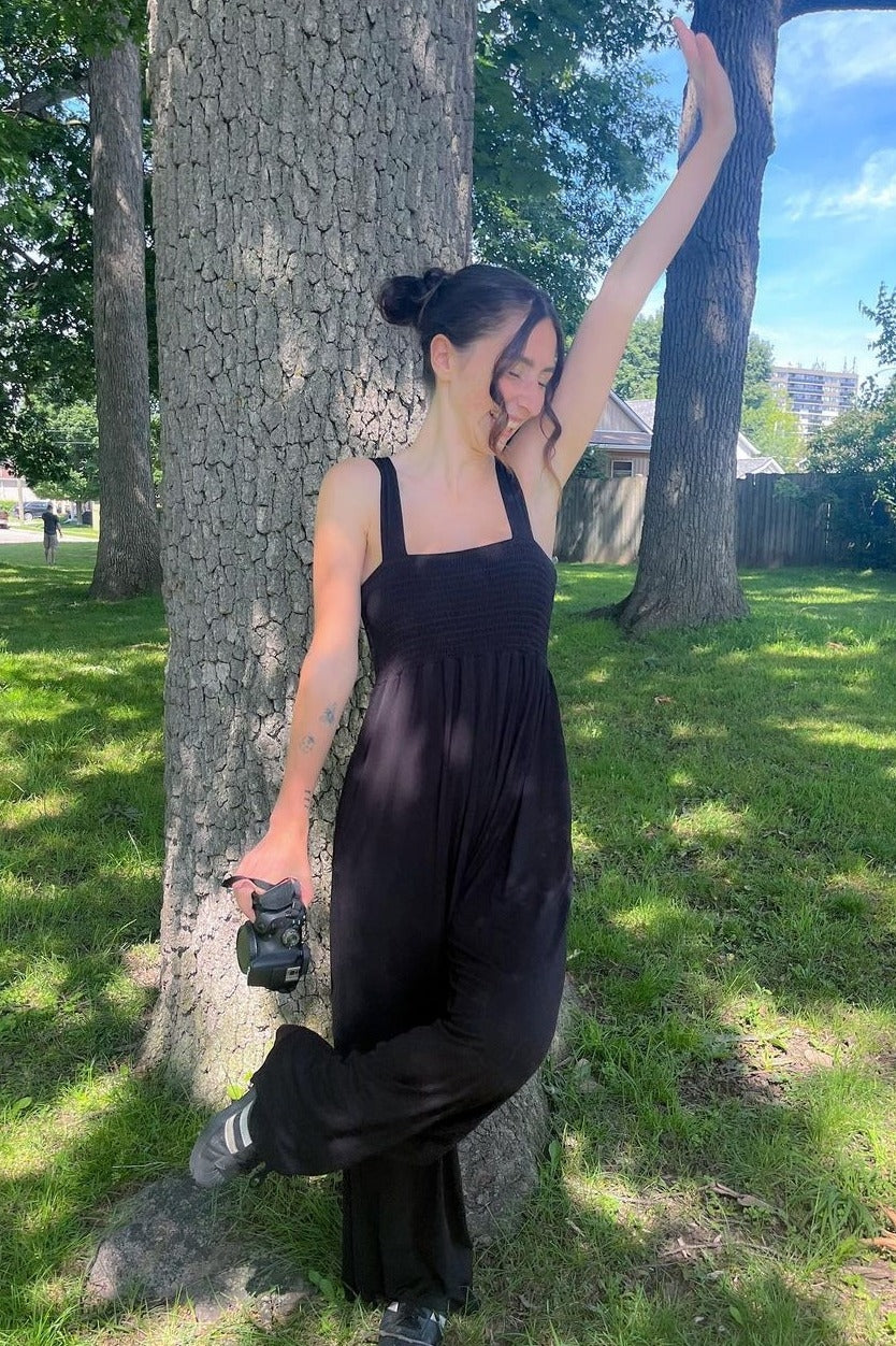 woman wearing a black smocked jumpsuit, standing by a tree and holding a camera while striking a pose