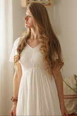 woman wearing a white short sleeve dress with a smocked bodice and flowing skirt, styled in a cozy indoor setting