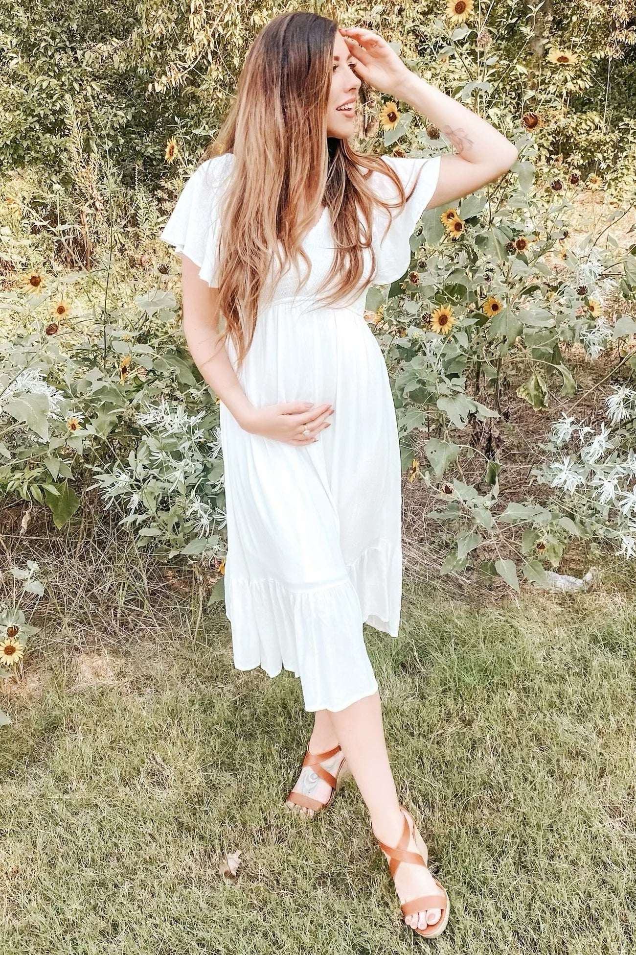 woman wearing a white maternity dress with flutter sleeves and a tiered hem, holding her baby bump in a garden