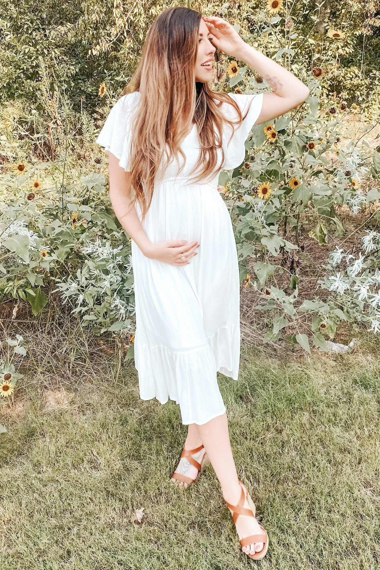woman wearing a white maternity dress with flutter sleeves and a tiered hem, holding her baby bump in a garden