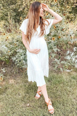 woman wearing a white maternity dress with flutter sleeves and a tiered hem, holding her baby bump in a garden