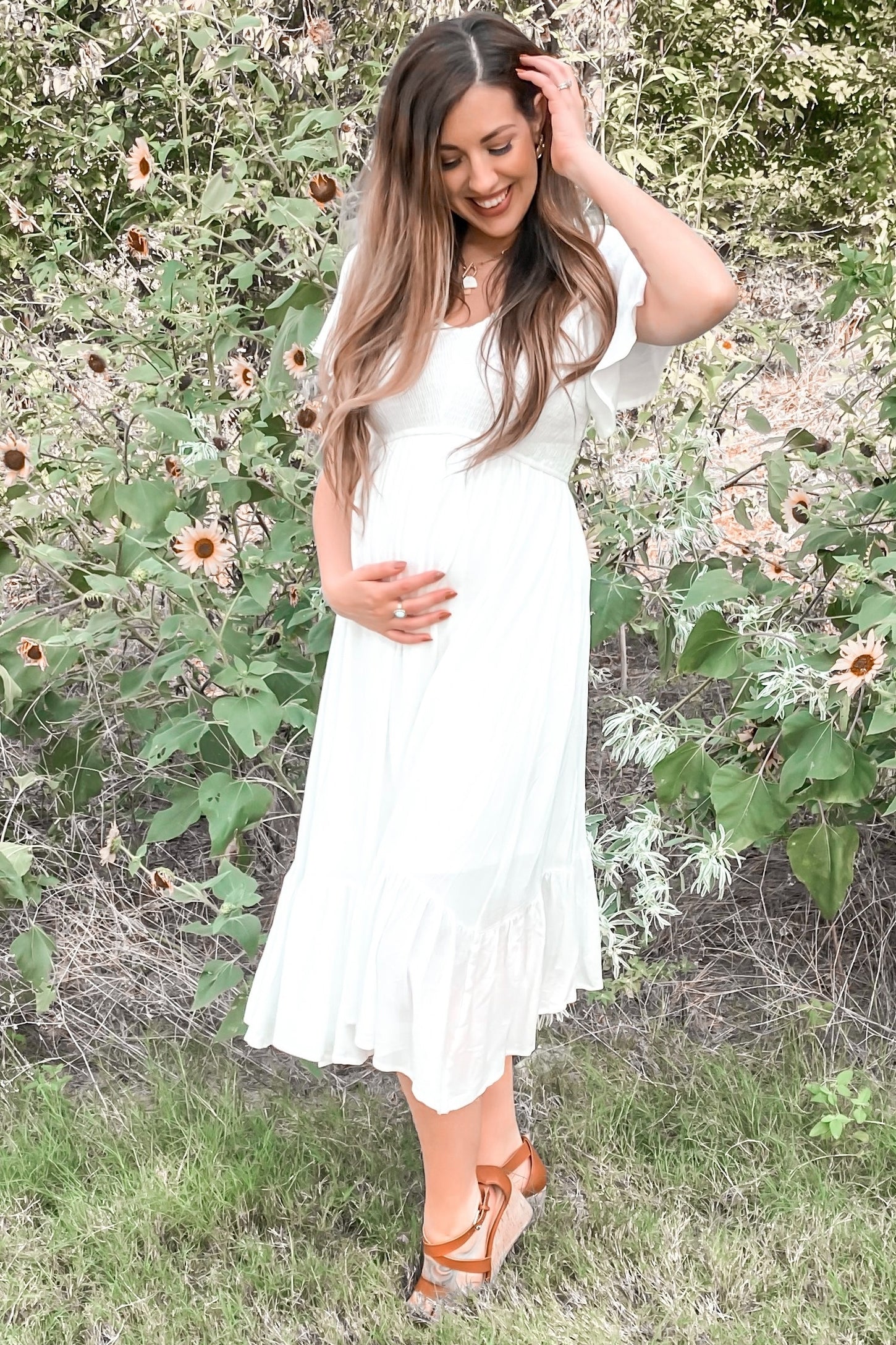 woman wearing a white maternity dress with flutter sleeves and a ruffled hem, gently holding her baby bump in a floral setting