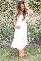 woman wearing a white maternity dress with flutter sleeves and a ruffled hem, gently holding her baby bump in a floral setting