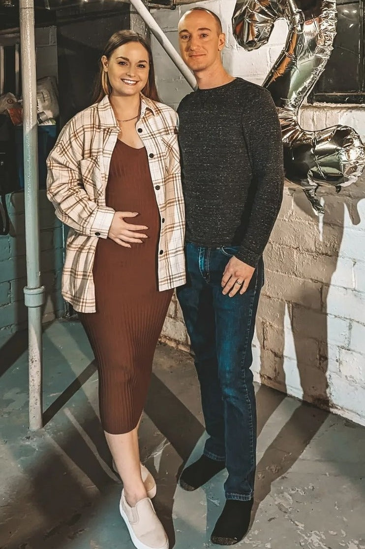 woman wearing a brown ribbed maternity dress and a plaid oversized shirt, cradling her baby bump with a partner beside her