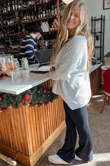 woman wearing a light gray oversized sweater and black flared pants, standing by a bar with festive decor