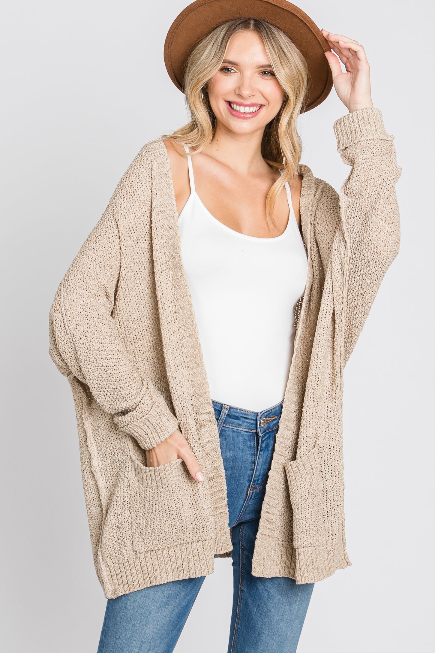 woman wearing a beige open knit cardigan with pockets over a white tank top and jeans, smiling and wearing a hat