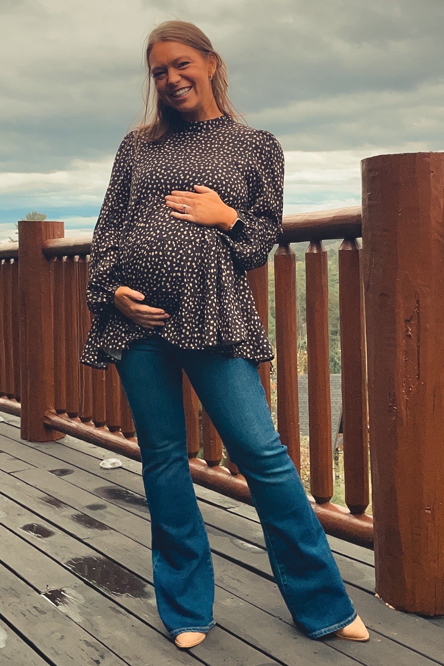 woman wearing a black polka dot maternity top and blue flared jeans, holding her baby bump outdoors