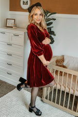 woman wearing a burgundy velvet maternity dress, cradling her baby bump, standing beside a crib and a teddy bear