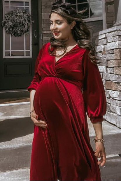 woman wearing a red velvet maternity dress and cradling her baby bump while standing outside a house