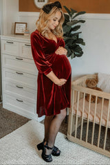 woman wearing a burgundy velvet maternity dress, holding her baby bump, with black shoes and a bow in her hair