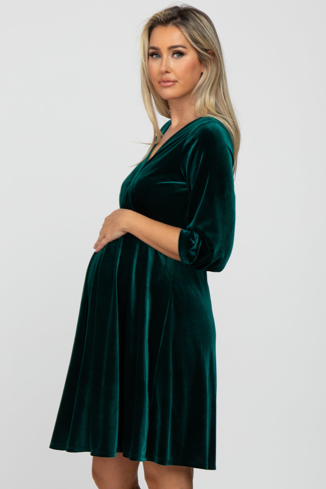 Pregnant woman wearing a dark green velvet maternity dress with three-quarter sleeves against a white background