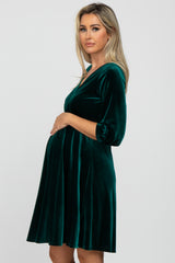 Pregnant woman wearing a dark green velvet maternity dress with three-quarter sleeves against a white background