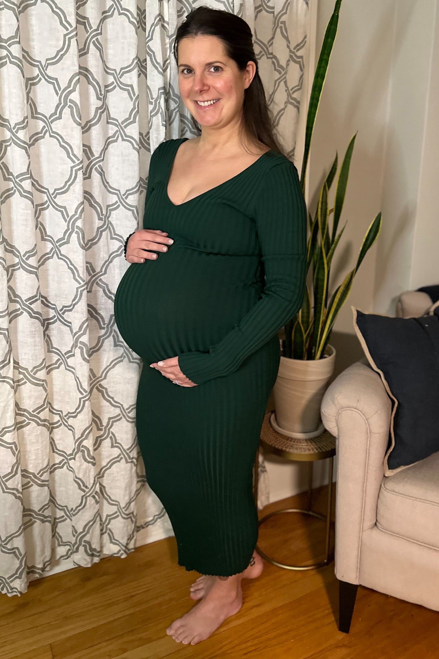 smiling pregnant woman in a dark green ribbed maternity dress holding her baby bump indoors