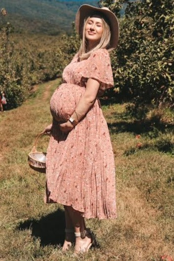 woman wearing a pink floral maternity dress with short sleeves and a baby bump, standing in an orchard with a basket