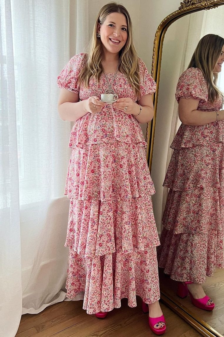 woman wearing a pink floral maternity dress with tiered ruffles, holding a cup, standing next to a mirror