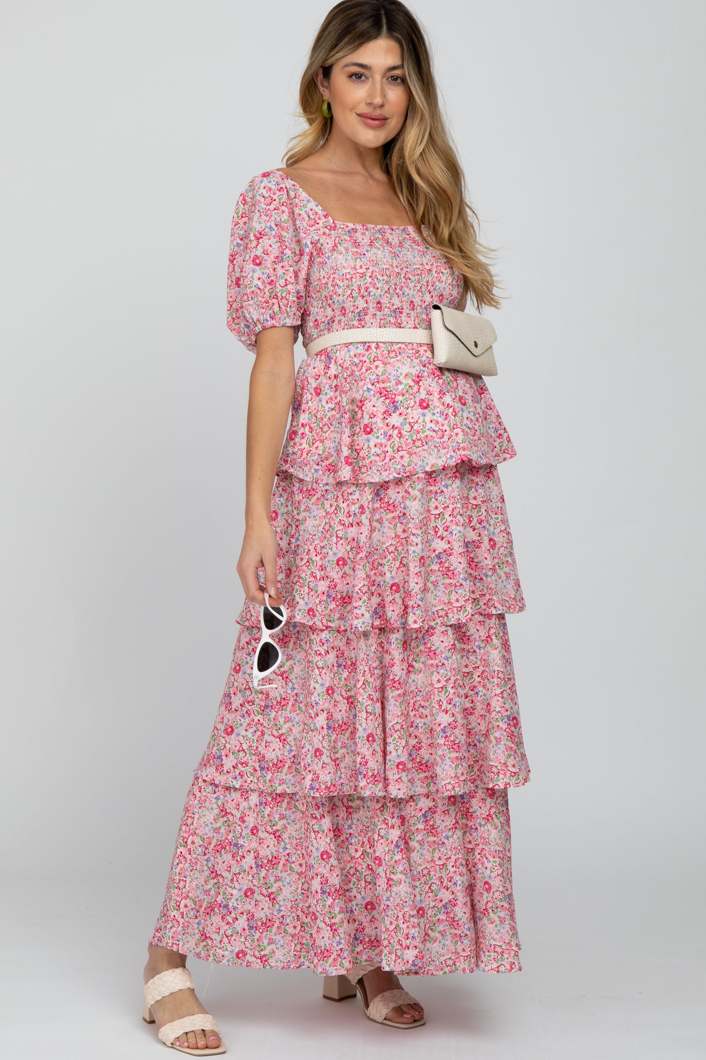 woman wearing a pink floral maternity dress with tiered ruffles and a white belt, styled with sunglasses and a small bag