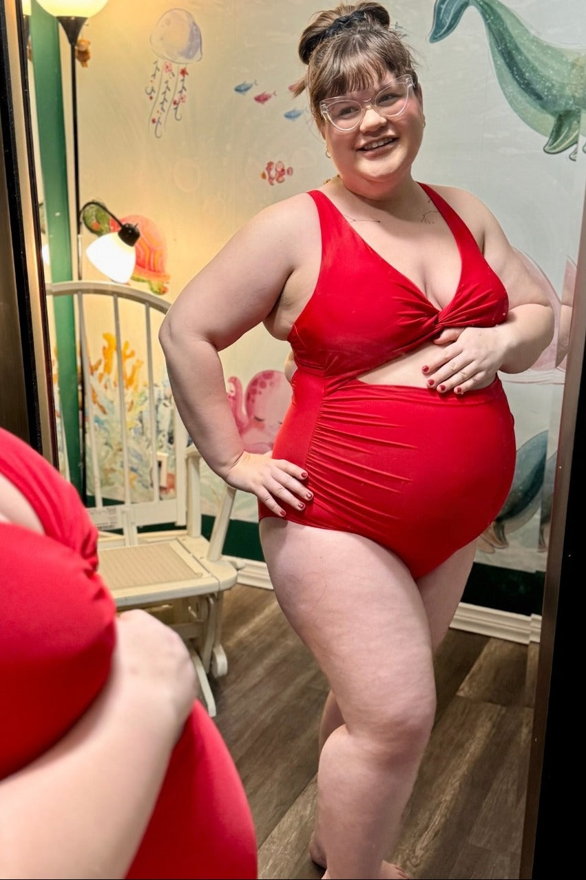 woman wearing a red maternity swimsuit with a twist front design and visible baby bump standing and smiling
