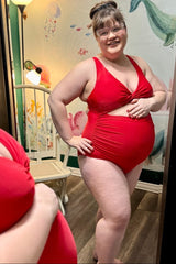 woman wearing a red maternity swimsuit with a twist front design and visible baby bump standing and smiling