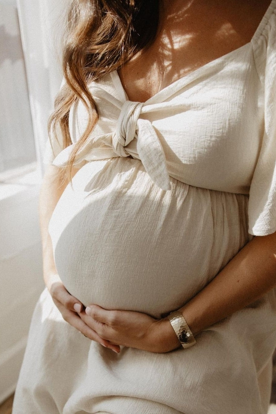 woman wearing a cream maternity dress with a knotted neckline, cradling her baby bump